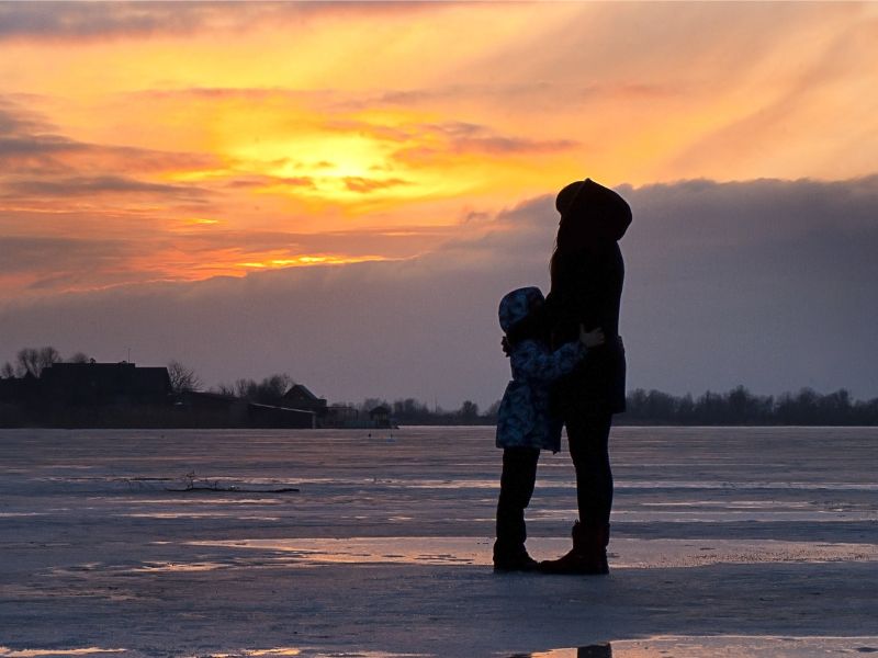 A silhouette of a mom and daughter standing on sand while watching a sun set.