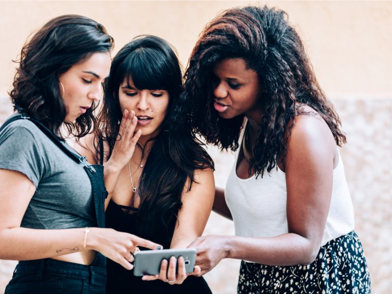 Three girls looking at a mobile phone, appearing to be surprised by the contents on the phone.