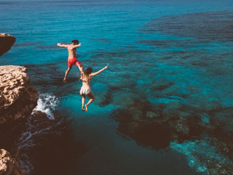 A man and woman jumping from a rock cliff into the blue ocean as an act of bravery.