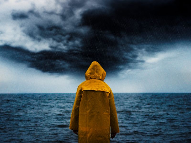 A person in a yellow rain jacket standing on a shoreline facing an oncoming storm.