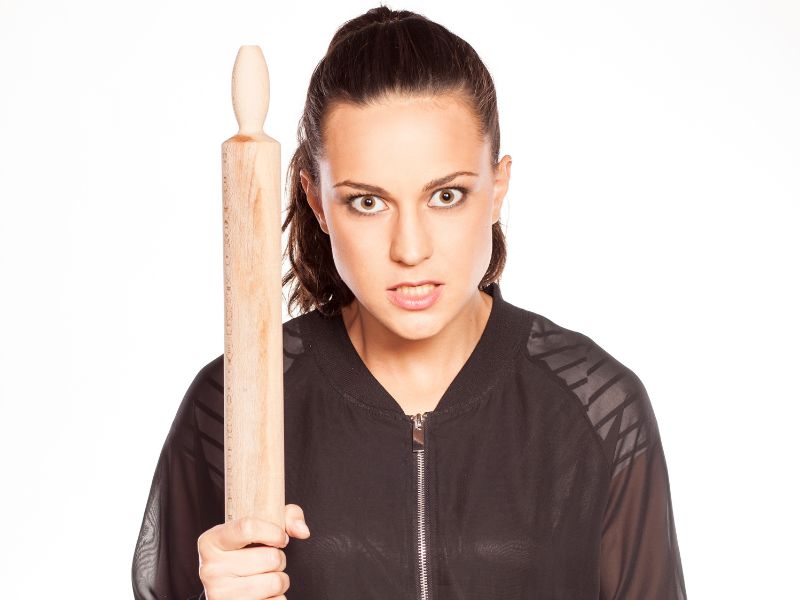 An angry woman wearing a black jacket with a hair tied back and holding a wooden utensil. 
