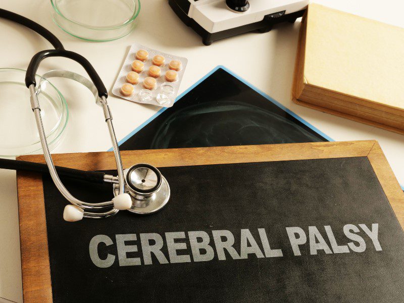 A black board with the words chalked ‘Cerebral palsy’ on a white table with a blister pack of orangey tablets, a doctor stethoscope, book and testing plate placed haphazardly around the board