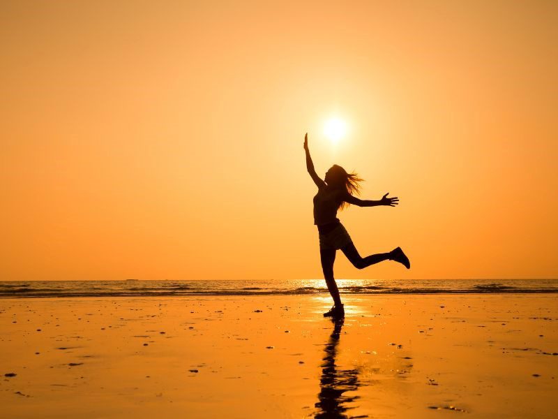 Image shows beautiful shades of peach beach sunset with the silhouette of a woman dancing with carefree abandon.