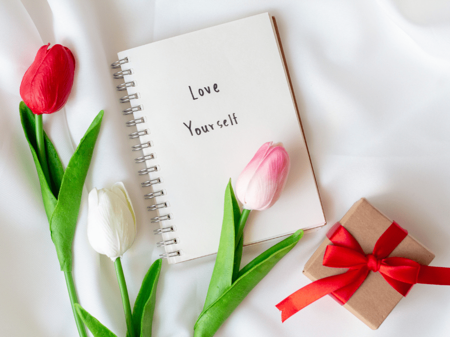 Image shows a spiral white notepad on a white sheet with the words, ‘love yourself’ written in black with 3 plastic flowers- one white, on in shades of pink and one cerise-thrown decoratively around it and a brown paper gift box with red ribbon wrapped into a bow.