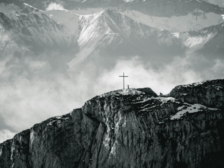 Image shows a cross in a mountain surrounded by snowy mountain ranges