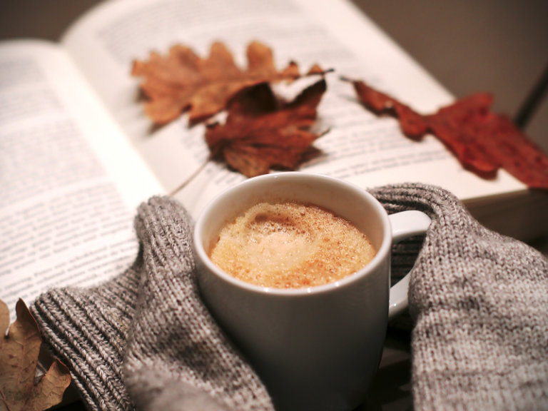 Warm cup of coffee beside an open book and cosy knitted blanket, representing relaxation, self-care and taking time for yourself.